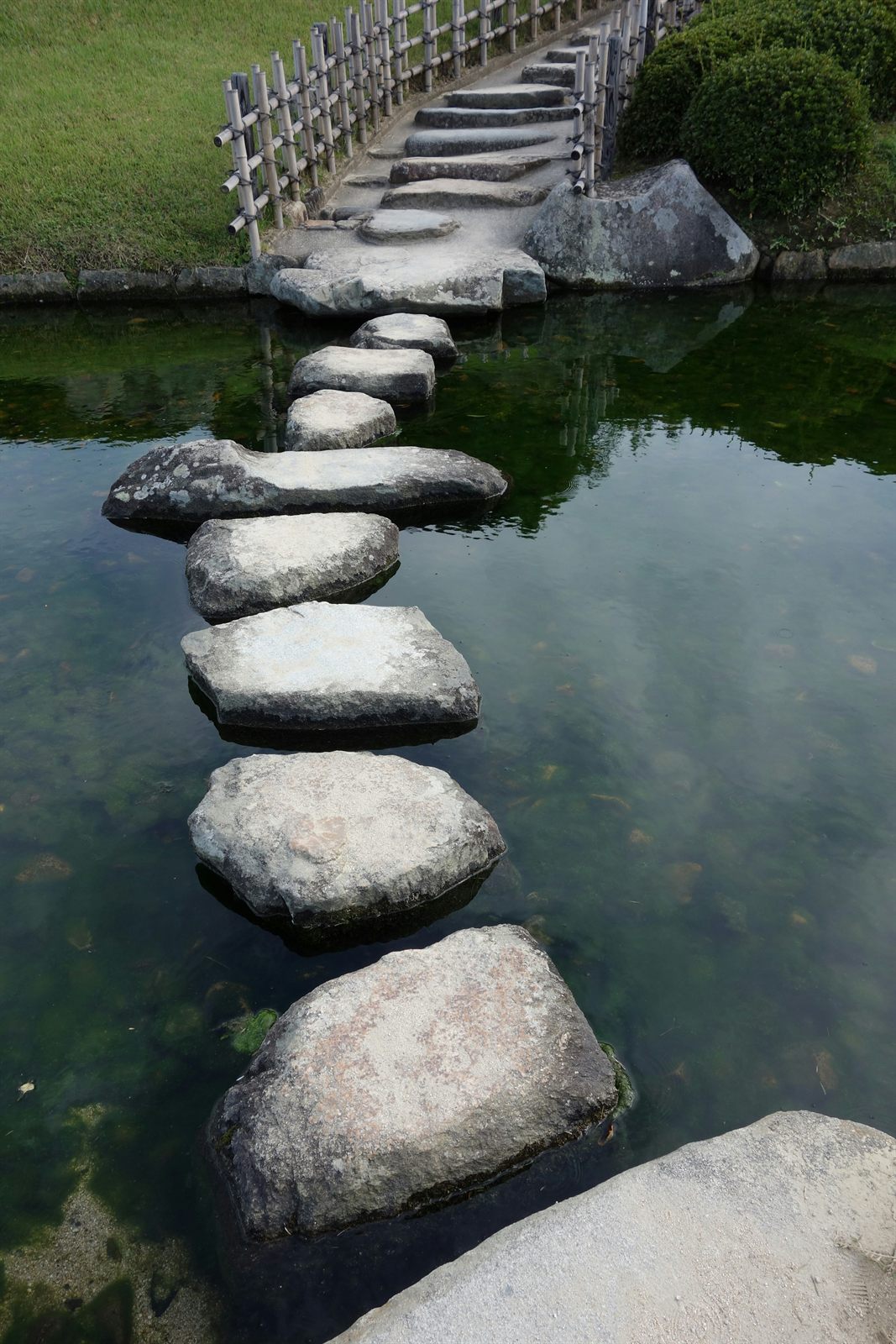 Stepping stones across a garden pond with a bamboo bridge beyond